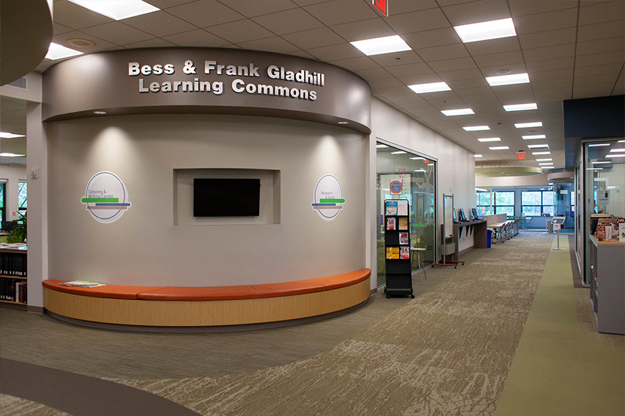 The main entrance to the Bess and Frank Gladhill Learning Commons, featuring a curved gray wall with a built-in orange bench and a flat-screen monitor. Wall graphics with arrows point left toward the Tutoring and Writing Center and right toward Research and Study areas.