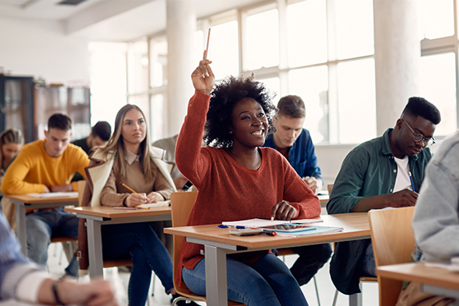 A confident student in a red sweater raises her hand in a bright classroom. Others are focused on their work, creating an atmosphere of active learning and engagement.